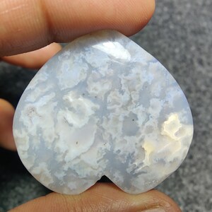 May include: A heart-shaped gemstone with a cloudy pattern of white and light blue. The stone is held between fingers, showcasing its polished surface and unique colour variations. The gemstone's design is a natural, organic shape.