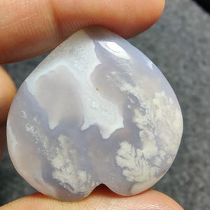 May include: A heart-shaped, polished gemstone with a light blue and white pattern. The stone has a smooth, glossy surface and a unique, natural design. The gemstone is held between fingers.