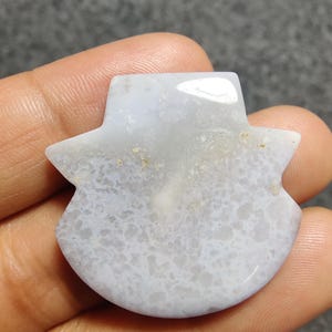 May include: A pale blue and white stone carving with a distinctive shape. The polished stone features a pattern of white and grey marbling. The carving is held in a hand, showing its size and detail. The stone appears to be a decorative item or a piece of jewellery.