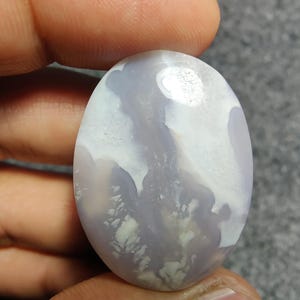 May include: An oval gemstone with a marbled appearance. The stone displays a blend of pale lavender and white colours, creating a cloudy effect. The polished surface reflects light, highlighting the natural patterns. A unique, natural stone, suitable for jewellery.