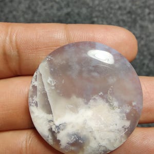 May include: A circular gemstone with a cloudy, layered design. The stone displays a blend of white, lavender, and grey tones, forming a natural, swirling pattern. The surface is smooth and reflective, with a slight sheen.