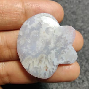 May include: A pale blue, heart-shaped gemstone with white and brown veining. The stone has a smooth, polished surface and a unique, organic shape. The gemstone is held in a hand.