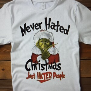 May include: White t-shirt with a graphic of a green character in a Santa-like outfit. The text reads "Never Hated Christmas Just Hated People" in black and red. The shirt is a crew neck style.