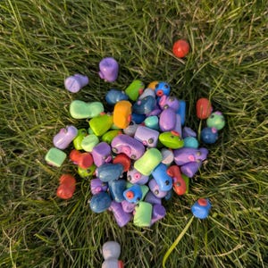 Puede incluir: Una colección de pequeñas figuras de arcilla hechas a mano, de colores brillantes, esparcidas sobre hierba verde. Las figuras son de varias formas y colores, incluyendo morado, azul, verde, naranja y rojo. Algunas tienen pequeños detalles.