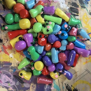 May include: A vibrant assortment of handmade clay figures in diverse shapes and sizes. The figures are predominantly in shades of red, yellow, green, blue, and purple, with black and pink details. They are arranged on a wooden surface with colourful paint splatters.