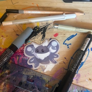 May include: A sticker of a cartoon moth with a white beard and crescent moon accents, surrounded by art supplies. A ruler, pencils, and markers are visible on a wooden surface with colorful paint splatters.