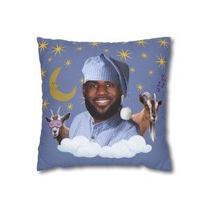 Puede incluir: Un cojín cuadrado con fondo azul. Presenta una persona sonriente con gorro de dormir y pijama a rayas, dos cabras, una luna creciente, estrellas y nubes. El diseño tiene un carácter caprichoso y onírico.