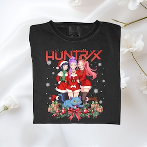 Puede incluir: Camiseta negra con un gráfico festivo de estilo anime. El diseño incluye tres personajes con trajes de Papá Noel, un gato azul y el texto rojo "HUNTRIX". Los copos de nieve y las decoraciones navideñas completan el tema festivo.