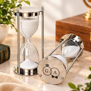 Könnte beinhalten: Zwei silberfarbene Sanduhren aus Metall mit klaren Glaskolben, die mit weißem Sand gefüllt sind. Eine Sanduhr steht aufrecht, während die andere auf der Seite liegt. Der Boden der liegenden Sanduhr zeigt eine schwarze Gravur von zwei ineinandergreifenden Ringen mit den Buchstaben G und A im Inneren, gekrönt von einem Diamantsymbol und einer Fliege.