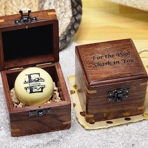 May include: Two dark brown wooden boxes with metal latches. One box is open, revealing a cream-colored billiard ball personalized with the letter E and the name Emilia. The closed box features the engraved text For the Pool Shark in You.