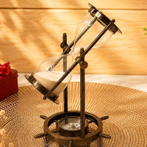 May include: A brass-toned hourglass with white sand, mounted on a rotating frame shaped like a ship's wheel. The base of the frame features an integrated circular compass. The hourglass is tilted at an angle, supported by vertical metal rods.