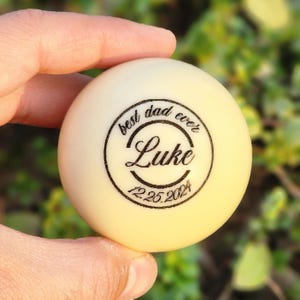 Puede incluir: Una pelota de ping pong de color crema con el texto "best dad ever" que rodea el nombre "Luke" y la fecha "12.25.2024" en tinta negra. La pelota se sostiene en una mano sobre un fondo verde borroso.