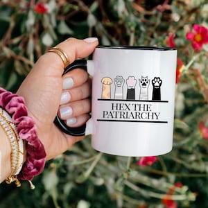 May include: White ceramic mug with a black handle and rim, featuring a row of stylized cat paw prints in various colors. The text "HEX THE PATRIARCHY" is printed below the paw prints in black.