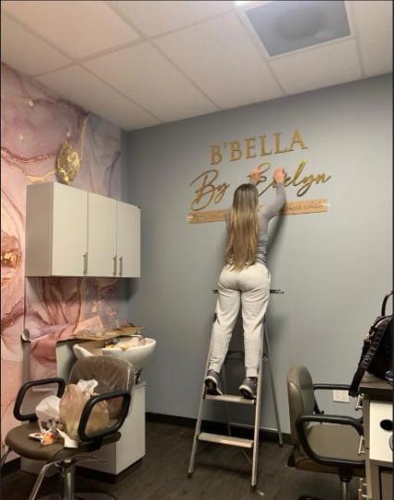 May include: A person on a step ladder installing a gold-colored sign that reads "B'BELLA By Evelyn" on a gray wall. The salon interior features a marble-patterned accent wall, a hair washing station, and salon chairs.