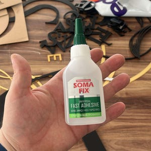 May include: A bottle of "Soma Fix" fast adhesive with a green nozzle. The label reads "Universal Fast Adhesive" and includes text in other languages. Black and gold letters are in the background.