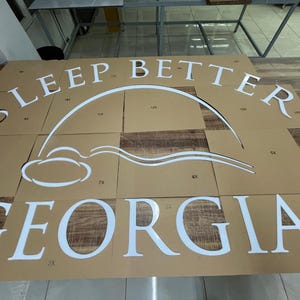 May include: White cut-out letters and design elements spelling "SLEEP BETTER GEORGIA" on a brown cardboard surface. The design includes a stylized cloud and arc. The letters are large and bold, suggesting a sign or decorative piece.