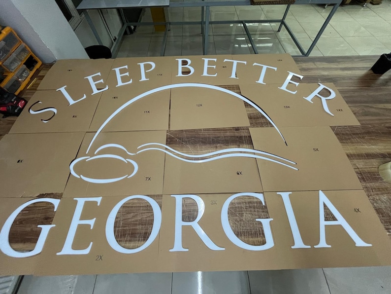 May include: White cut-out letters and design elements spelling "SLEEP BETTER GEORGIA" on a brown cardboard surface. The design includes a stylized cloud and arc. The letters are large and bold, suggesting a sign or decorative piece.