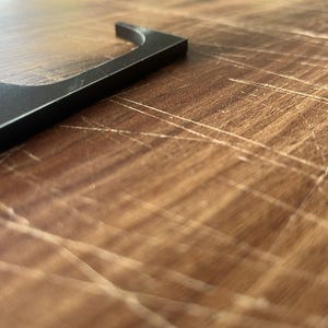 May include: A black metal letter "L" rests on a wooden surface. The letter is in the upper left corner, with the rest of the image showing the wood grain. The wood has a warm, brown tone with visible scratches.