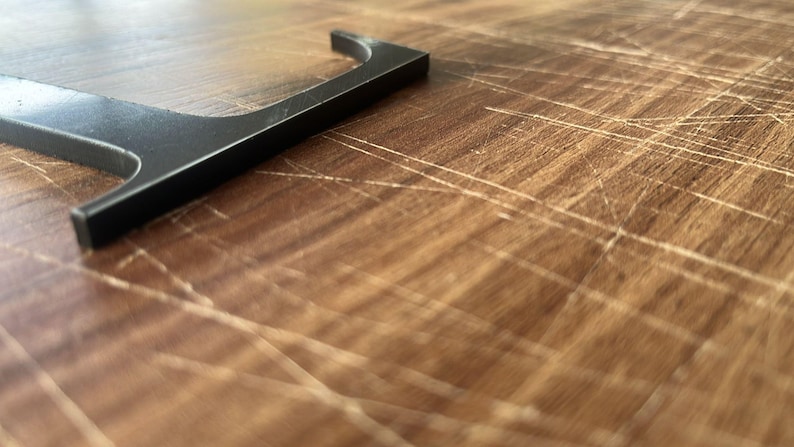 May include: A black metal letter "L" rests on a wooden surface. The letter is in the upper left corner, with the rest of the image showing the wood grain. The wood has a warm, brown tone with visible scratches.