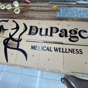 May include: A partially assembled sign for "DuPage Medical Wellness" with black lettering and a stylized graphic. The sign is on a tan surface, with the words "MEDICAL WELLNESS" below "DuPage".
