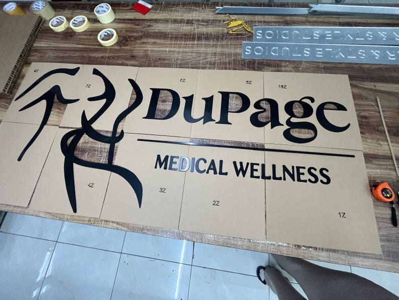 May include: A partially assembled sign for "DuPage Medical Wellness" with black lettering and a stylized graphic. The sign is on a tan surface, with the words "MEDICAL WELLNESS" below "DuPage".