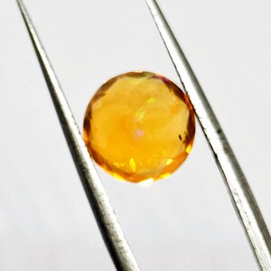 May include: A faceted, round, golden-yellow gemstone is held by silver tweezers against a white background. The gem has a brilliant, multi-faceted cut, reflecting light and displaying a warm, radiant glow. The tweezers are positioned diagonally.
