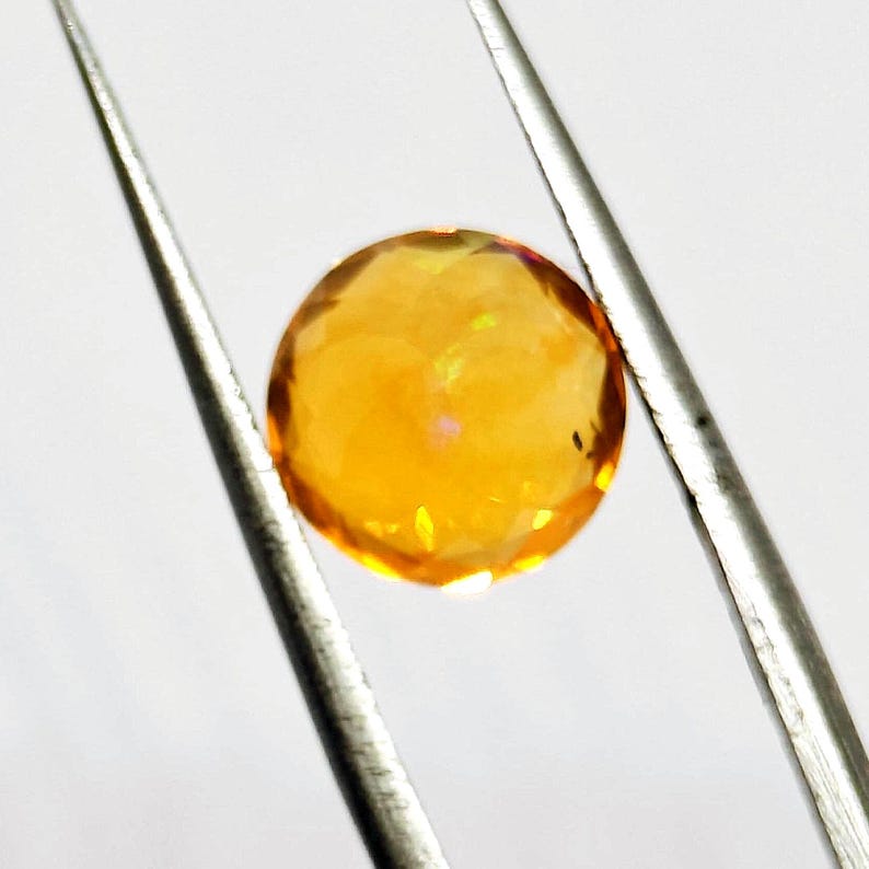 May include: A faceted, round, golden-yellow gemstone is held by silver tweezers against a white background. The gem has a brilliant, multi-faceted cut, reflecting light and displaying a warm, radiant glow. The tweezers are positioned diagonally.