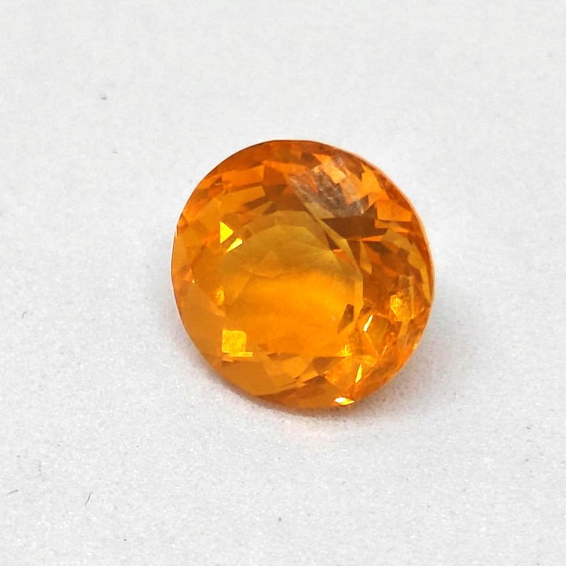 May include: A faceted, round-cut gemstone with a vibrant orange hue. The gem's surface reflects light, showcasing its multiple facets. The gemstone is set against a white background, highlighting its color and clarity. This gemstone is suitable for jewelry making.