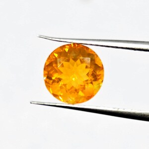May include: A round, faceted gemstone held by tweezers. The gem is a vibrant orange color, with a brilliant, star-like pattern visible in its center. The gemstone is set against a plain white background.