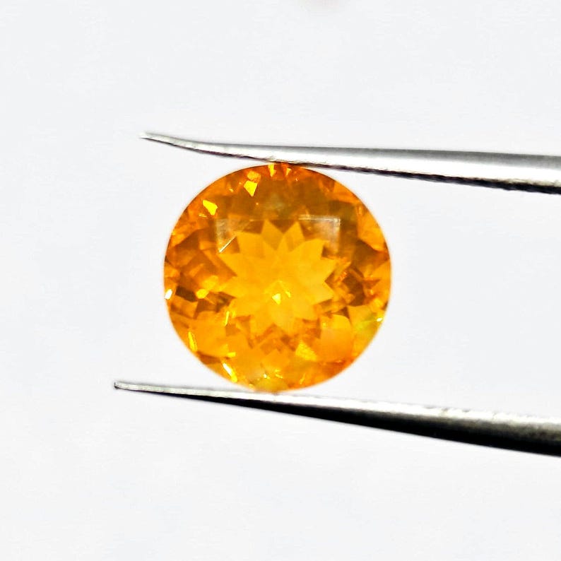 May include: A round, faceted gemstone held by tweezers. The gem is a vibrant orange color, with a brilliant, star-like pattern visible in its center. The gemstone is set against a plain white background.