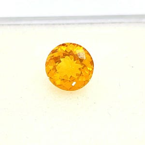 May include: A faceted, round, golden-yellow gemstone. The gem has a brilliant cut, with numerous small facets creating a sparkling effect. The stone is set against a white background, highlighting its color and clarity. This gemstone is suitable for jewelry.