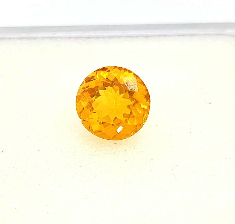 May include: A faceted, round, golden-yellow gemstone. The gem has a brilliant cut, with numerous small facets creating a sparkling effect. The stone is set against a white background, highlighting its color and clarity. This gemstone is suitable for jewelry.