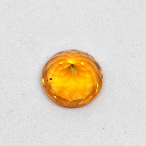 May include: A faceted, round gemstone with a warm, golden-orange hue. The gem has a brilliant cut, reflecting light across its many facets. The stone is set against a white background, highlighting its color and clarity.