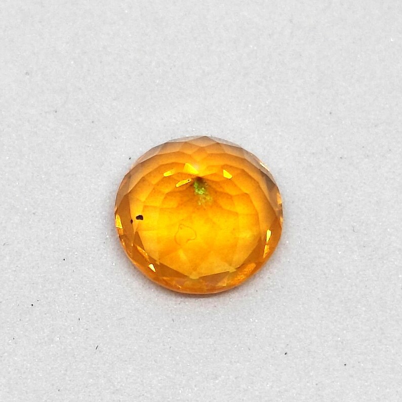 May include: A faceted, round gemstone with a warm, golden-orange hue. The gem has a brilliant cut, reflecting light across its many facets. The stone is set against a white background, highlighting its color and clarity.