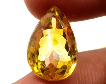 May include: A faceted, pear-shaped gemstone with a vibrant yellow-orange hue. The gem is held between fingers, showcasing its polished surfaces and internal clarity. The cut of the stone creates multiple reflective facets.
