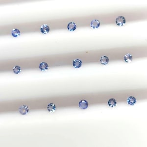 May include: A selection of small, round, light blue gemstones displayed on a white surface. The gemstones have a faceted appearance, reflecting light. The arrangement is in rows, with varying sizes of the gemstones.