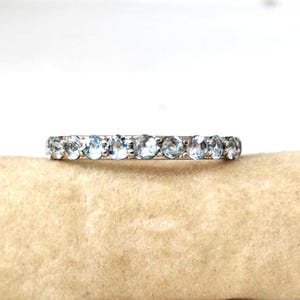 May include: A silver ring with a continuous row of round, light blue gemstones. The ring is set against a neutral background, highlighting the sparkle of the gemstones. The ring is a delicate piece of jewellery.