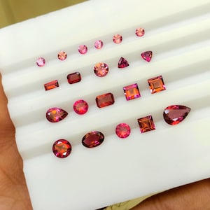 May include: An assortment of faceted gemstones in various shapes and shades of pink and red. The gemstones are displayed on a white tray, showcasing their different cuts, including round, oval, square, and pear shapes. The gems vary in size.