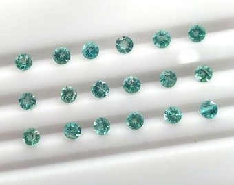 May include: A collection of round, teal-colored gemstones arranged on a white surface. The gemstones are faceted, reflecting light and showcasing their color. The arrangement suggests a display of loose gemstones.