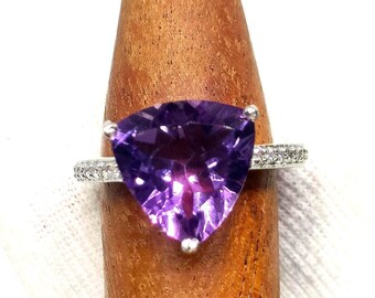 May include: A silver ring featuring a large, triangular, deep purple amethyst gemstone. The band is adorned with small, clear gemstones. The ring is displayed on a wooden ring holder against a white background.