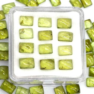 Puede incluir: Una colección de piedras preciosas de peridoto de corte cuadrado, verde claro, exhibidas en una caja de plástico transparente. Las piedras son translúcidas, con otras piedras de peridoto sueltas.