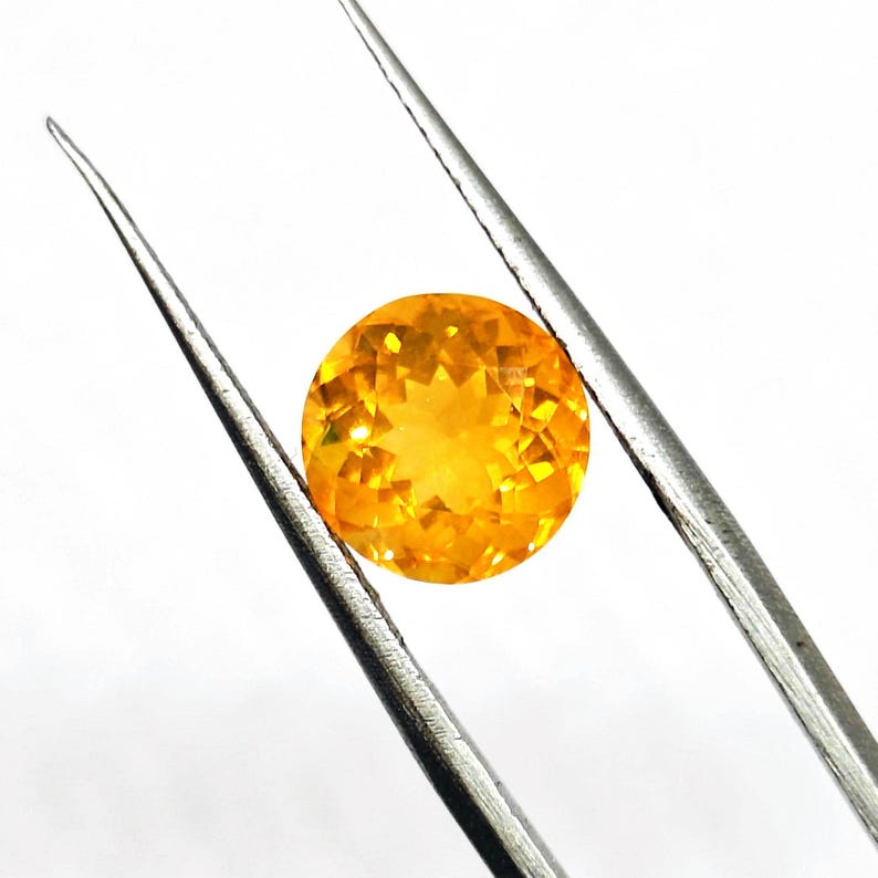 May include: A round, faceted, golden-yellow gemstone is held by silver tweezers against a white background. The gemstone has a brilliant cut, reflecting light and showcasing its clarity and color. The tweezers are positioned diagonally.
