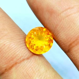 May include: A round, faceted gemstone with a vibrant yellow-orange hue. The gem is held between two fingers, showcasing its brilliant cut and color. The gemstone appears to be a citrine or similar type of precious stone.