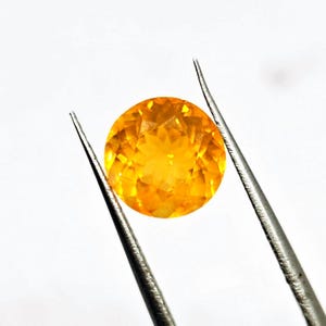 May include: A round, faceted gemstone with a vibrant yellow-orange hue, held by silver tweezers against a white background. The gemstone's cut reveals multiple facets, creating a sparkling effect. The gem appears to be approximately 0.75 inches in diameter.