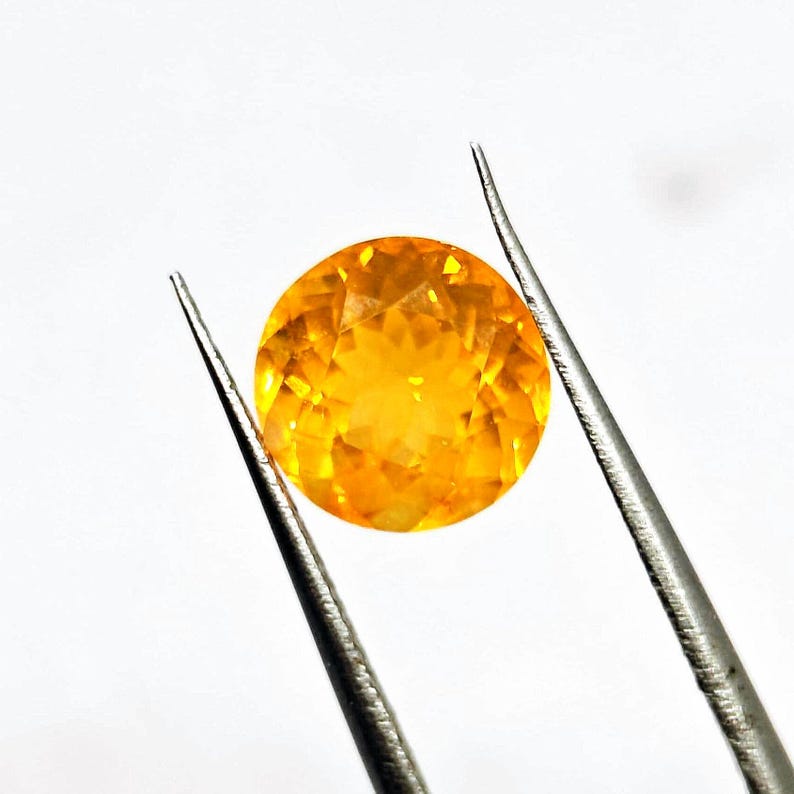 May include: A round, faceted gemstone with a vibrant yellow-orange hue, held by silver tweezers against a white background. The gemstone's cut reveals multiple facets, creating a sparkling effect. The gem appears to be approximately 0.75 inches in diameter.