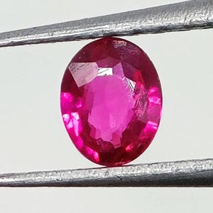 Puede incluir: Una gema de rubí ovalada y facetada sujeta con pinzas metálicas. El rubí es de un color rojo rosado intenso y vibrante, con inclusiones internas visibles. La gema se encuentra sobre un fondo blanco liso.
