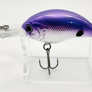 May include: A fishing lure with a metallic purple back that fades into a silver, diamond-patterned side. It features a clear plastic diving lip, a black eye, a black spot on the side, and two silver treble hooks attached to the underside and tail.