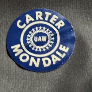 May include: A circular blue badge featuring the white text "CARTER MONDALE" around the edge. The centre has a white design and the letters "UAW". The badge is set against a grey background.