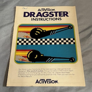 Puede incluir: Instrucciones del videojuego Dragster de Activision. La portada presenta un diseño retro con un gráfico de coche de carreras negro, una bandera a cuadros y el título del juego. El texto dice "Dragster Instructions" y "Activision."