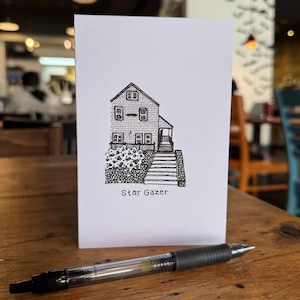 May include: A white greeting card with a black ink illustration of a house with the text "Star Gazer". A black and silver pen rests on a wooden surface in front of the card.